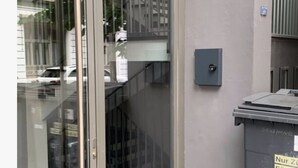 Ein Gebäudeeingang mit Glastür, Metalltreppe und Mülleimer.