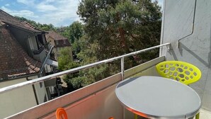 A balcony with a white table and two chairs, one orange and one yellow, overlooking a residential area with trees and houses. - Cozy, sunny, city center 1BR flat (Marble 3) (Zürich)
