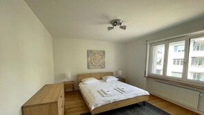 Slaapkamer met een bed, houten nachtkastjes, een plafondventilator, een radiator en een raam met uitzicht op gebouwen.