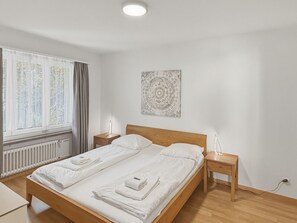 Una habitación de hotel moderna y luminosa, con cama de madera, ropa de cama blanca, una mesita de noche con lámpara y una ventana con cortinas.