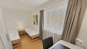 A compact hotel room with two single beds, a desk with a chair, a small bedside table with a lamp, and a window with curtains. - Beautiful, quite and very central 2BR flat (Sky6) (Zürich)