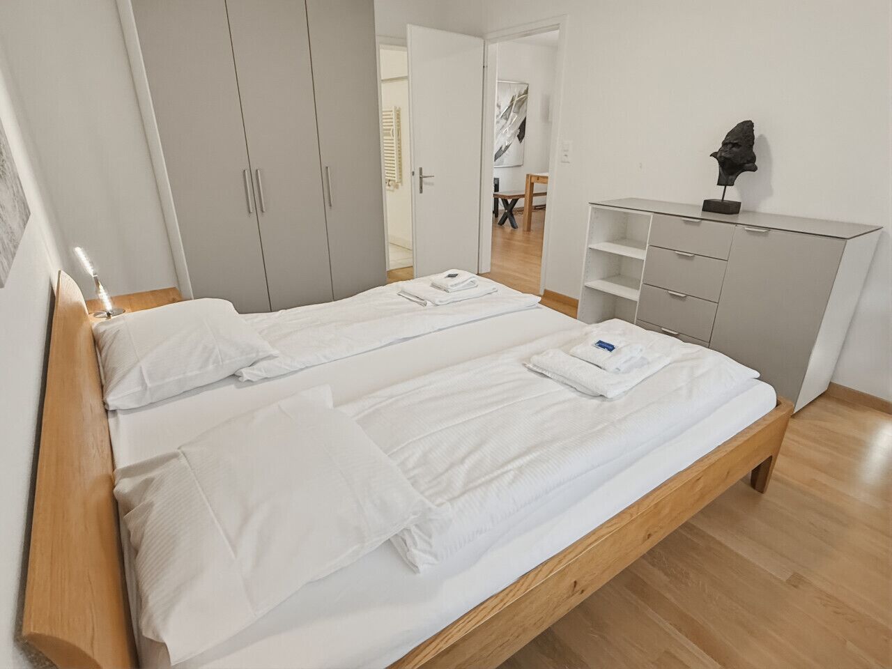 A bedroom with a wooden bed, white bedding, a grey wardrobe, and a desk with a sculpture.