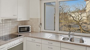 A kitchen with white cabinets, a microwave, a sink, and a window with a view of trees. - Beautiful, quite and very central 2BR flat (Sky6) (Zürich)