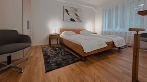 A bedroom with a wooden bed, a nightstand, a chair, and a window with sheer curtains. - Fantastic 2BR in the city center (Sky7) (Zürich)
