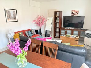 Een woonkamer met een bank, een eettafel met een bloevas, een televisie en een boekenkast.