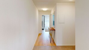 Een smalle gang met witte wanden en houten vloer, die leidt naar een kamer met een deur en een klein tafeltje.