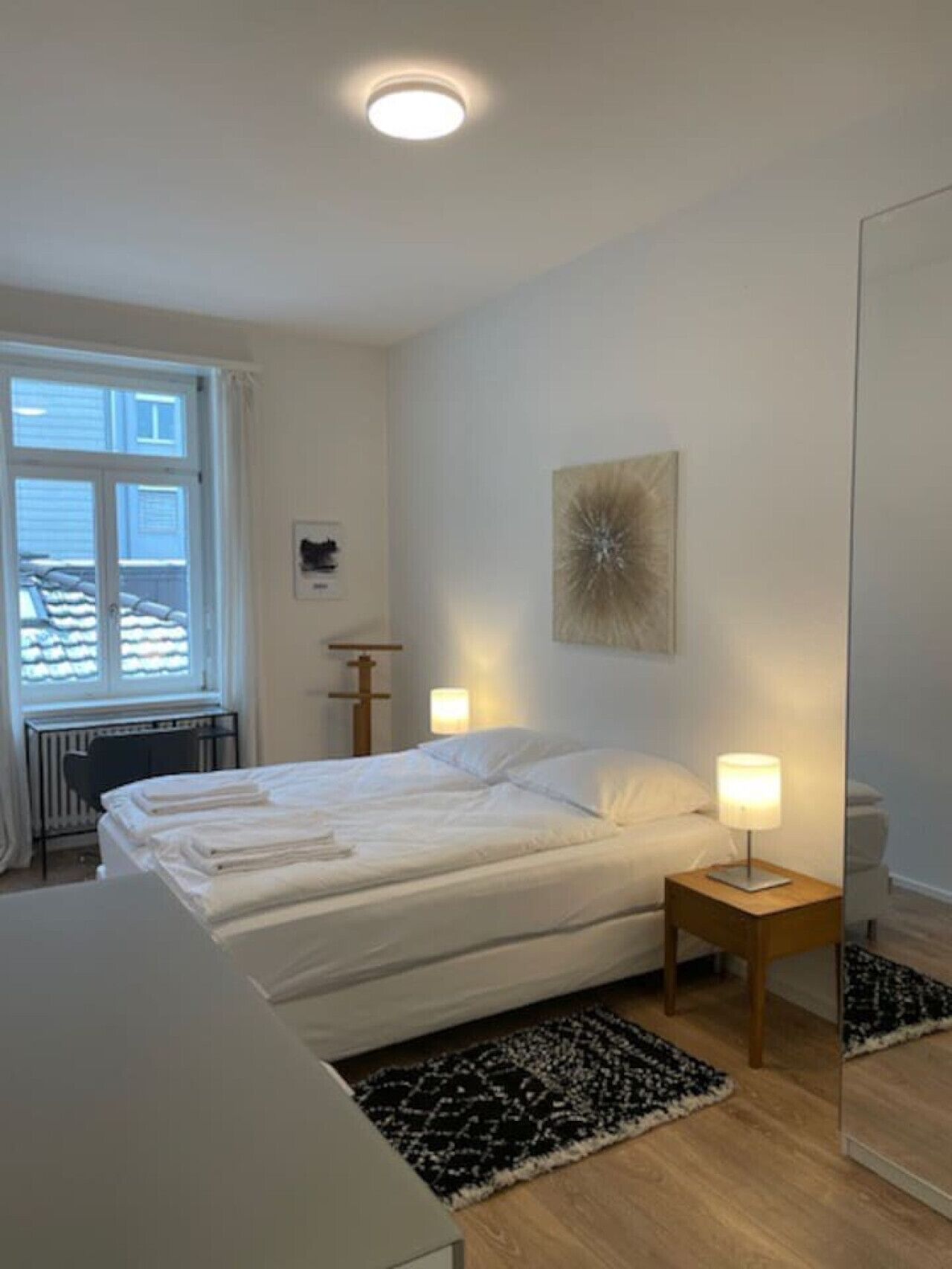 Een slaapkamer met een bed, nachtlampjes, een radiator, een raam en een aan de muur bevestigd kunstwerk.