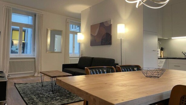 Ett modernt vardagsrum med ett matbord i trä, en svart soffa och en modern lampa.