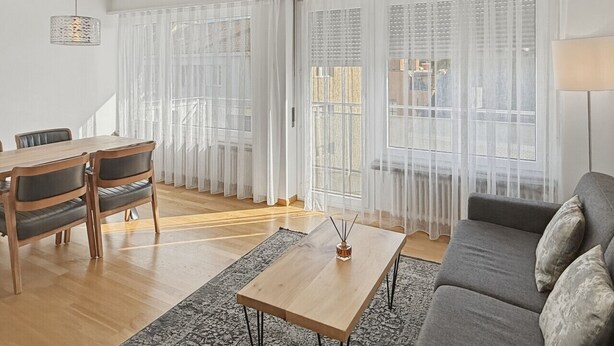 A modern living room with a dining area, large windows with sheer curtains, a grey sofa, and a wooden coffee table.