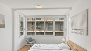 A bedroom with a wooden bed, two bedside lamps, a gray chair, and a large window with a view of buildings. - Amazing 2BR flat: spacious and central (Duke2) (Zürich)