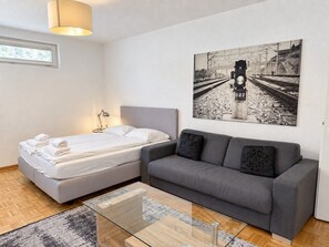 A modern bedroom with a bed, a sofa, a glass coffee table, and a wall-mounted picture of a railway track.