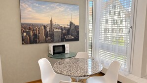 Un coin repas avec une table ronde, des chaises blanches, un four à micro-ondes et un tableau représentant un paysage urbain accroché au mur.