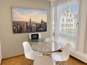 A dining area with a round table, white chairs, a microwave, and a cityscape painting on the wall.