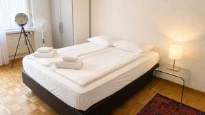 A neatly made bed with white linens and towels, a bedside lamp, and a floor lamp near a window with curtains. - Terrific studio in the city centre (Art 9) (Zürich)