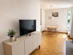 A modern living room with a flat-screen TV on a cabinet, a dining area with a table and chairs, and a large window allowing natural light.