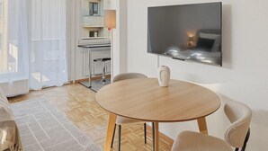 Een moderne woonkamer met een ronde houten tafel, twee comfortabele fauteuils, een flatscreen televisie aan de muur en een groot raam met dunne gordijnen.