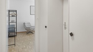 Een gang met een visgraatvloer, een witte deur, een kapstok met een kledingstuk eraan en een stoel.