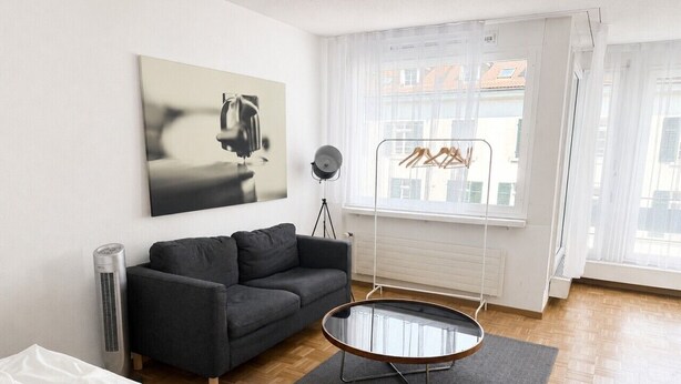 A modern living room with a sofa, a glass coffee table, a standing heater, and a window with a view of buildings. - Cozy studio in the city centre (Art 13) (Zürich)