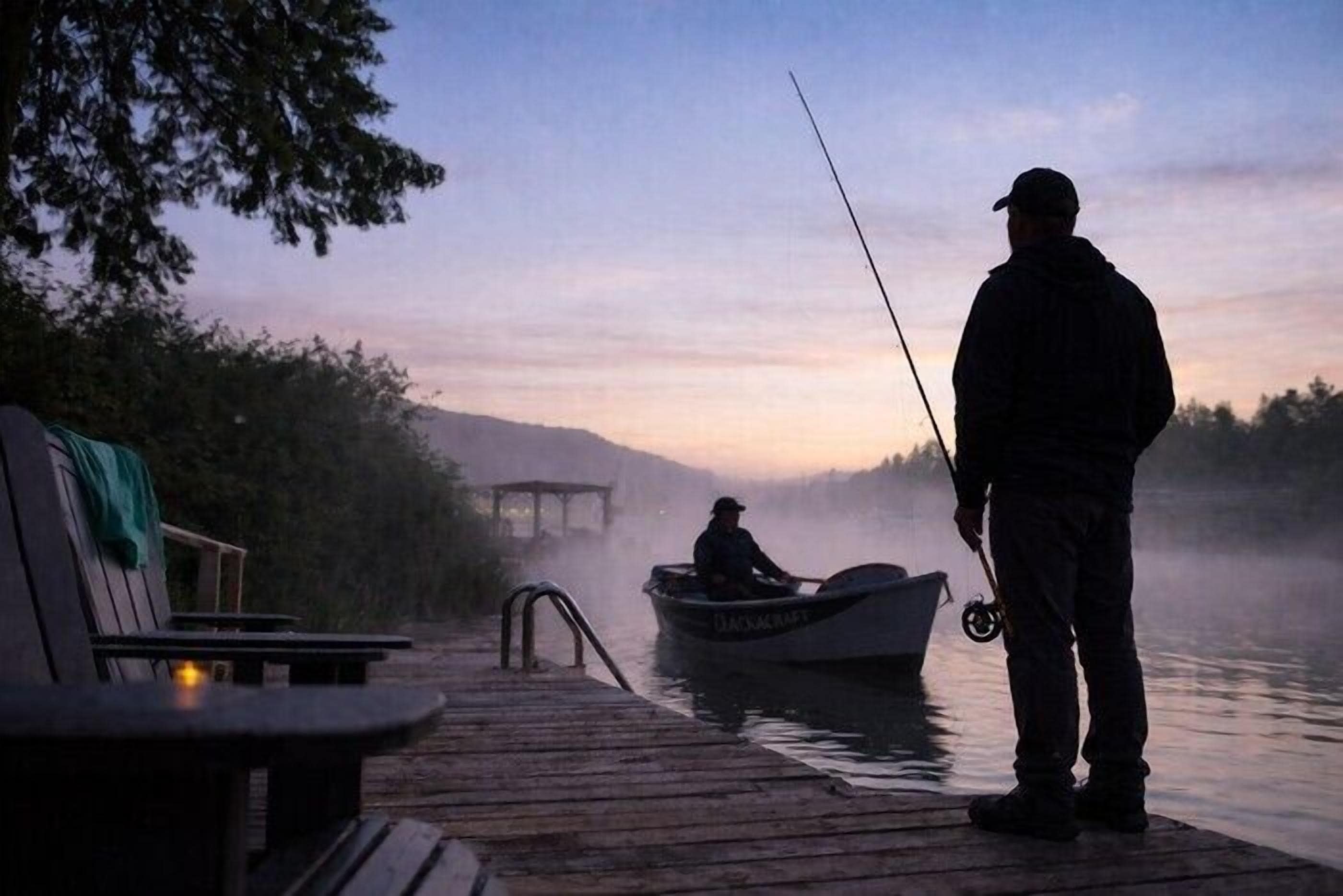 Un muelle de madera se adentra en un lago brumoso al amanecer o al atardecer, con una persona pescando y un pequeño bote cerca.