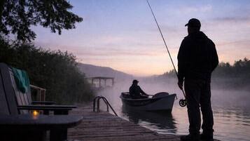 Un muelle de madera se adentra en un lago brumoso al amanecer o al atardecer, con una persona pescando y un pequeño bote cerca.