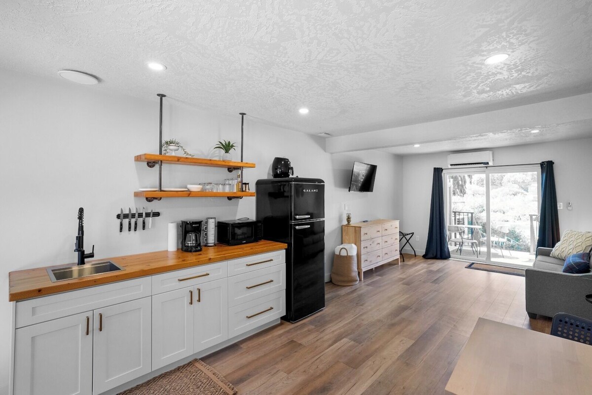A modern kitchen with white cabinets, a black refrigerator, and wooden countertops. There is a dining area with a gray sofa and a table.