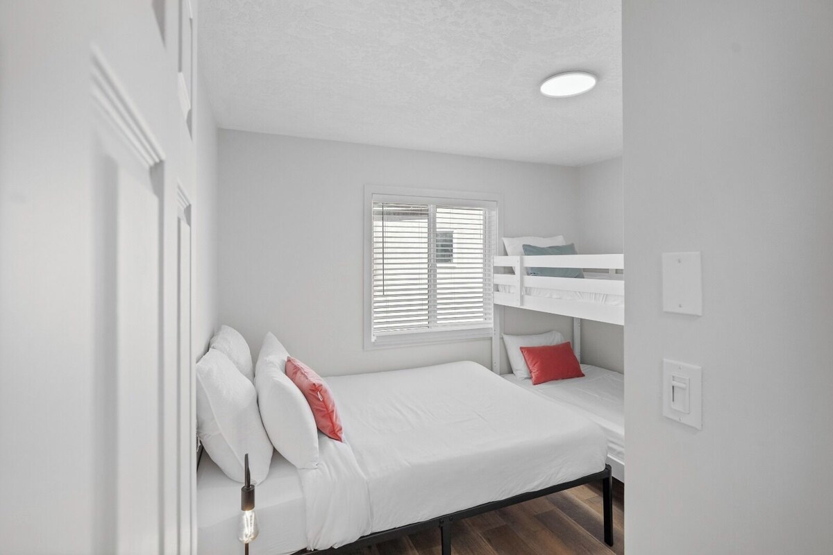 A room with a bunk bed, a single bed, white walls, a window with blinds, and a ceiling light.