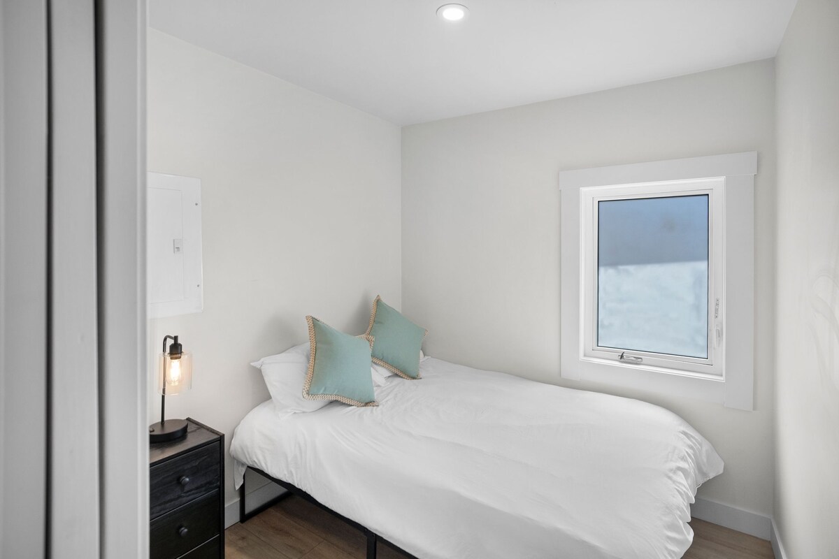 A bedroom with a bed, two pillows, a nightstand with a lamp, a window with a view, and a door.