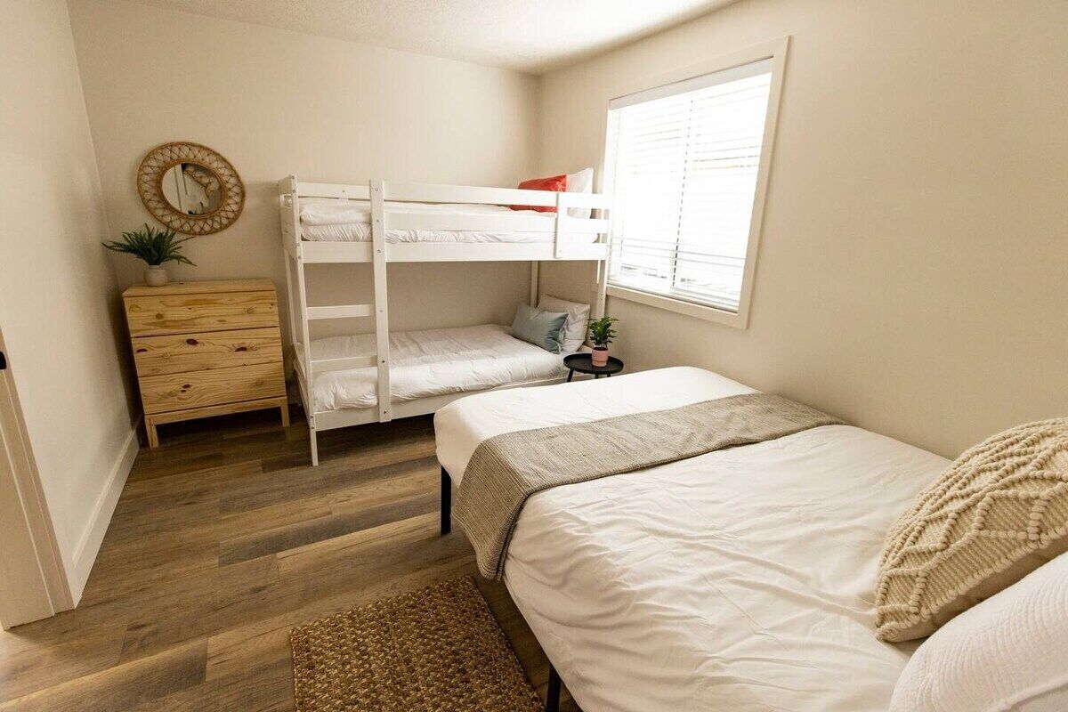 Un dormitorio con una cama litera, un aparador de madera, una mesita con una planta y una ventana con persianas.