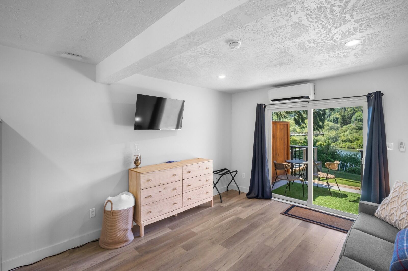 A modern room with a wooden floor, a flat-screen TV mounted on the wall, a wooden dresser, and a sliding glass door leading to an outdoor area with a view of greenery.
