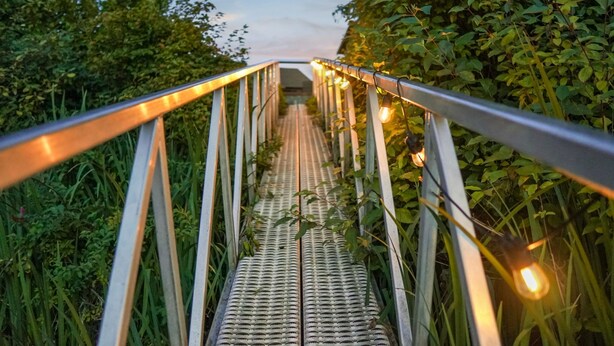 Un puente metálico con barandilla de metal y luces colgantes, rodeado de follaje.