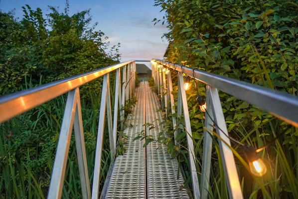 A metal bridge with a metal railing and string lights, surrounded by greenery. - Riverside by Cowichan Lake Cottages (Lake Cowichan)