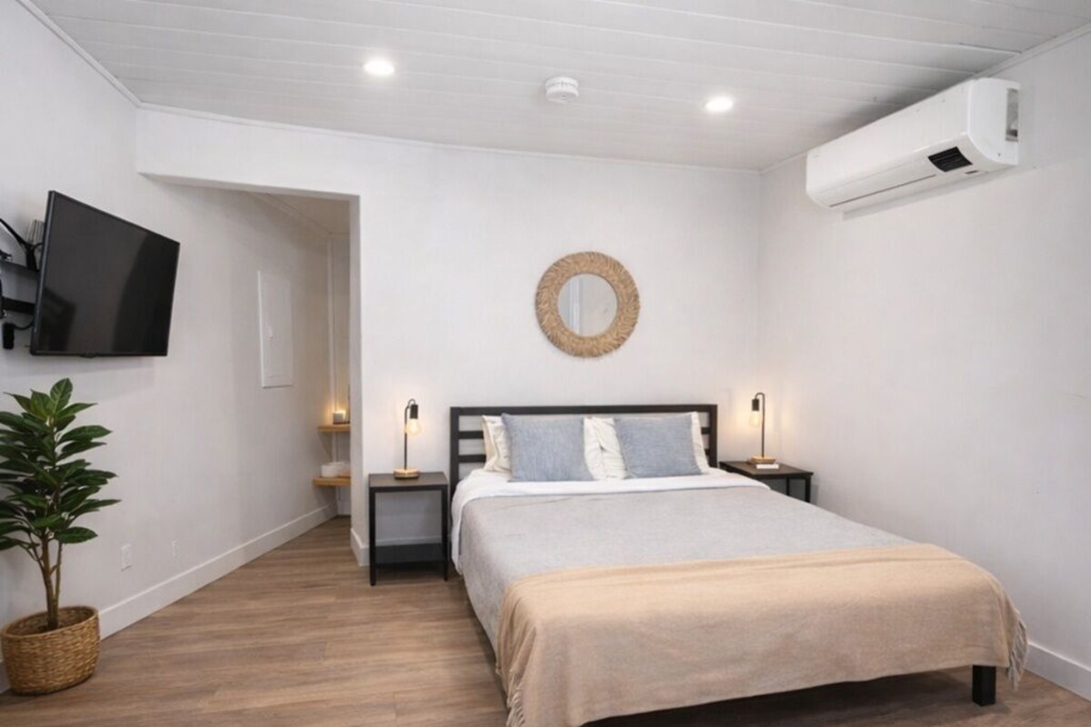A bedroom with a bed, bedside tables, a wall-mounted TV, a circular mirror, and an air conditioning unit.