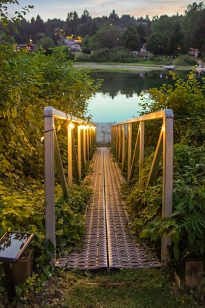 A wooden bridge with railings and lights, leading to a body of water surrounded by greenery. - Riverside by Cowichan Lake Cottages (Lake Cowichan)