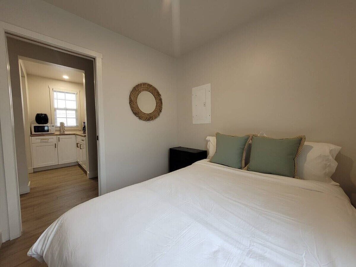 A bedroom with a large bed, a bedside table, and a wall-mounted circular decoration. A kitchen area is visible through an open doorway.