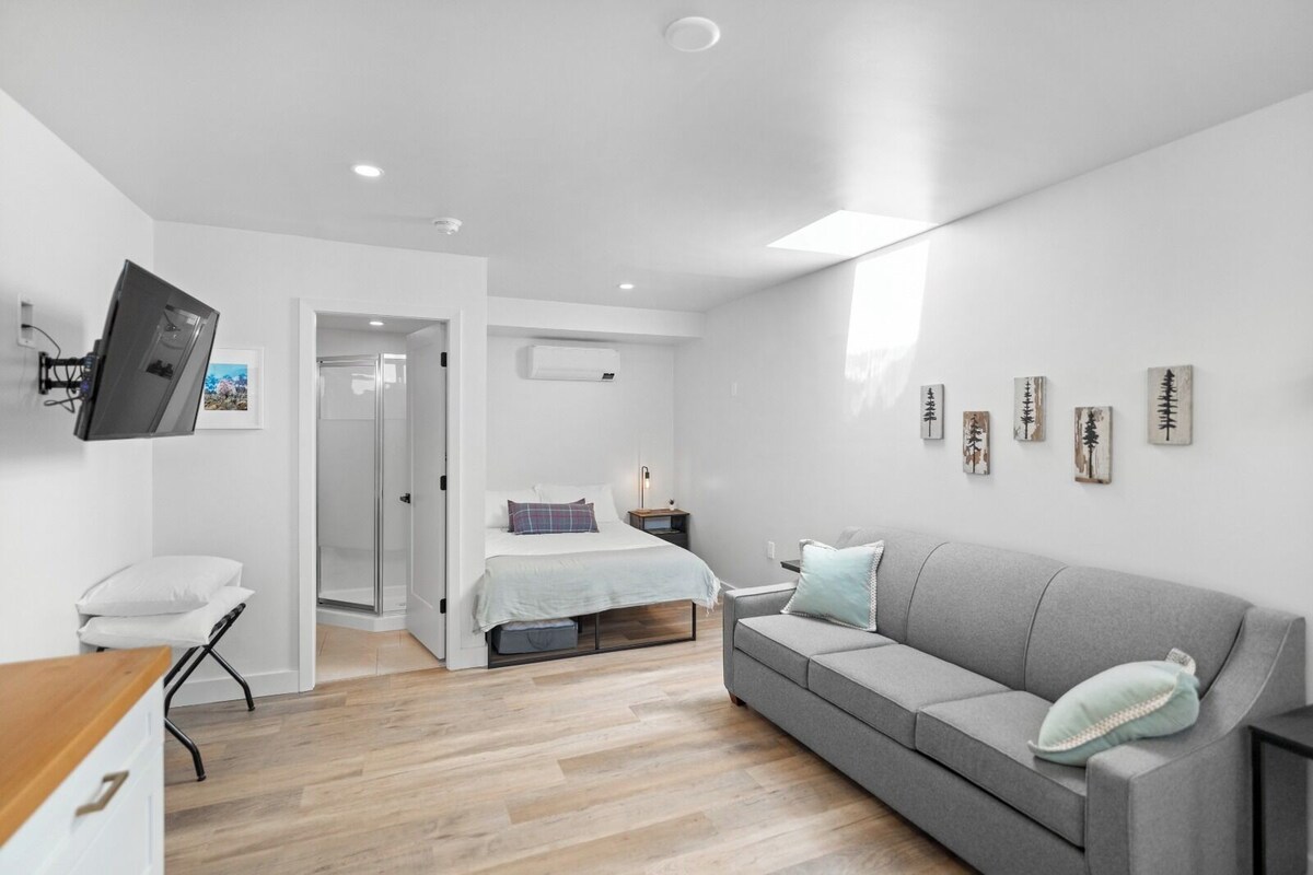 A modern bedroom with a bed, a grey sofa, a TV mounted on the wall, and a wooden floor.