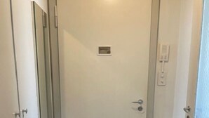Un couloir étroit avec une porte blanche, une petite grille d’aération et un interphone fixé au mur.