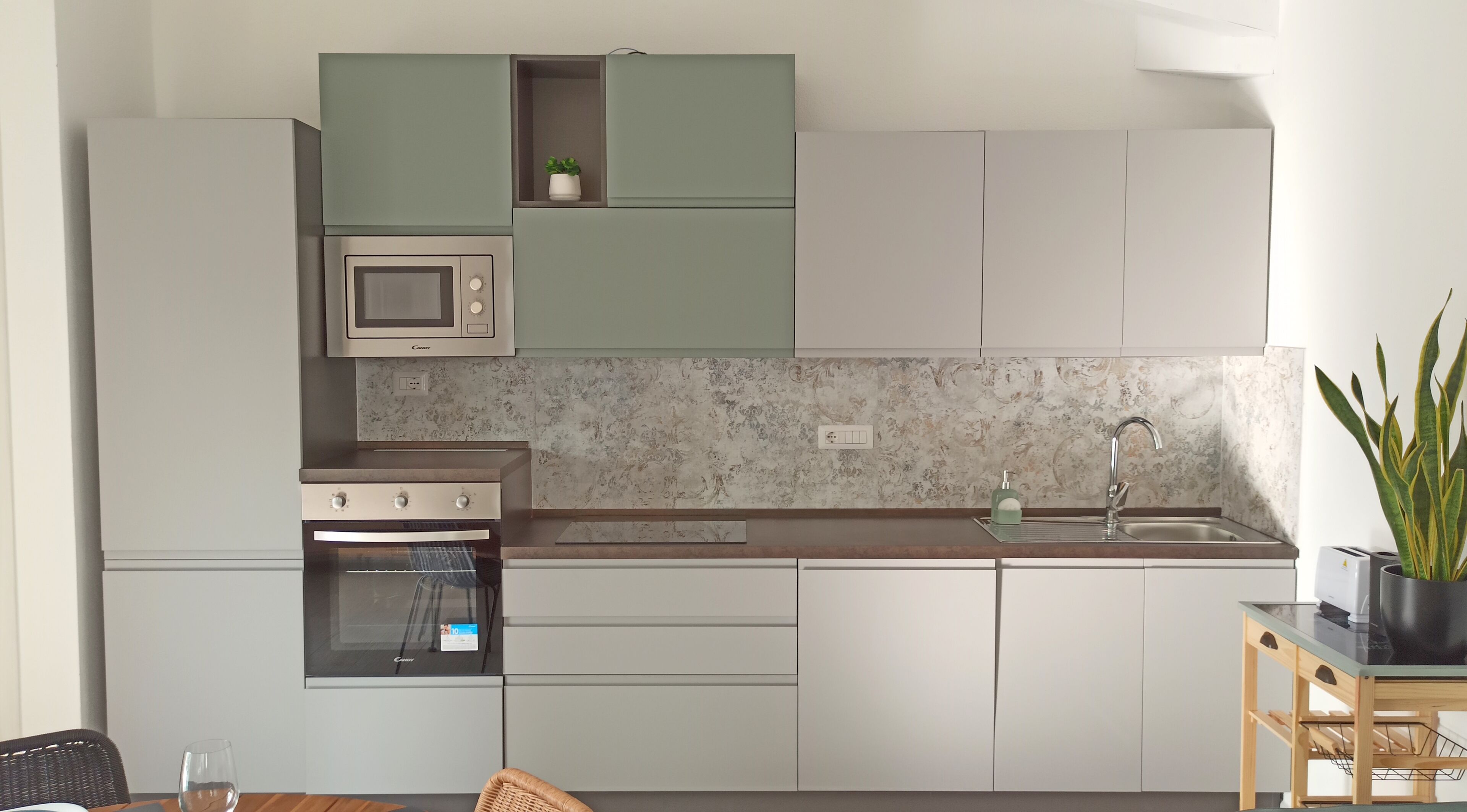 A compact kitchen with a built-in microwave, oven, and sink. There is a small plant on the countertop and a wicker chair in the foreground.
