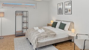 A bedroom with a bed, bedside tables, a chair, a floor lamp, a shelf with towels, and a window with blinds. - Charming studio in the city centre (Art 21) (Zürich)