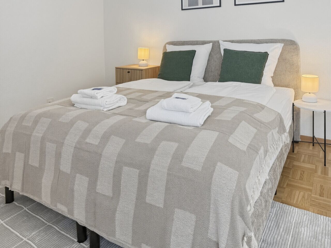 A neatly made bed with a checkered bedspread, two pillows, and folded towels on a wooden floor.