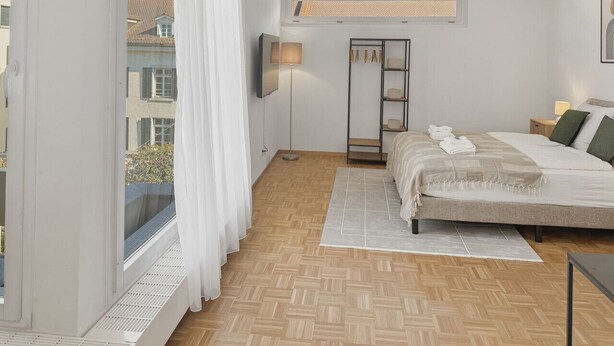 A bedroom with a bed, a bedside table, a lamp, a wardrobe, and a window with curtains. - Charming studio in the city centre (Art 21) (Zürich)