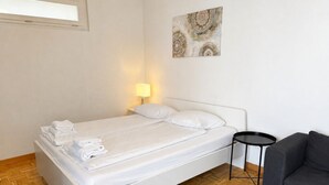 A neatly made bed with folded towels, a bedside lamp, a small round table, and a grey sofa in a room with wooden flooring and a white wall. - Practical studio in the city center (Art 7) (Zürich)