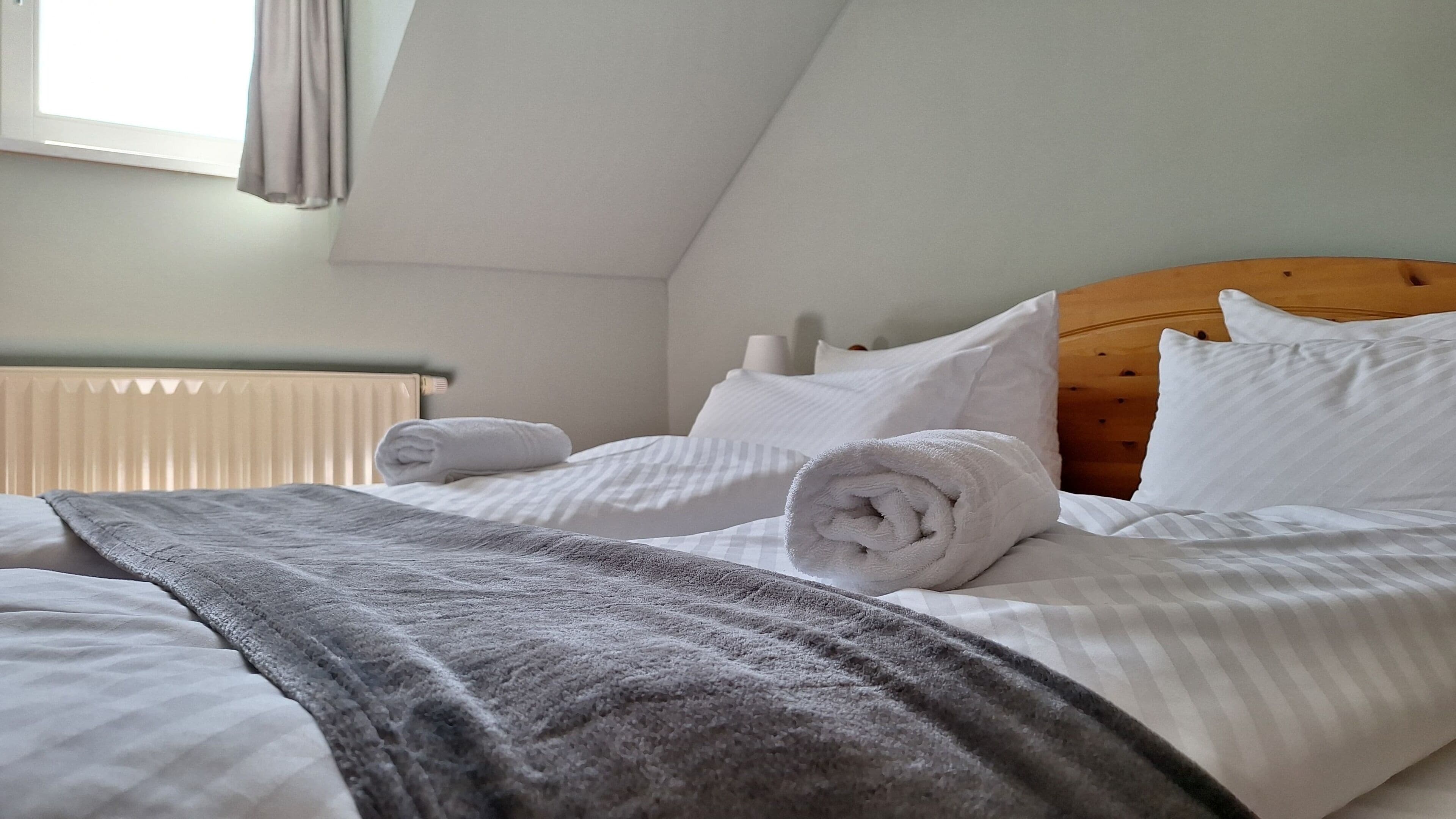 A neatly made bed with white linens and a grey blanket, a wooden headboard, and a radiator in the background.