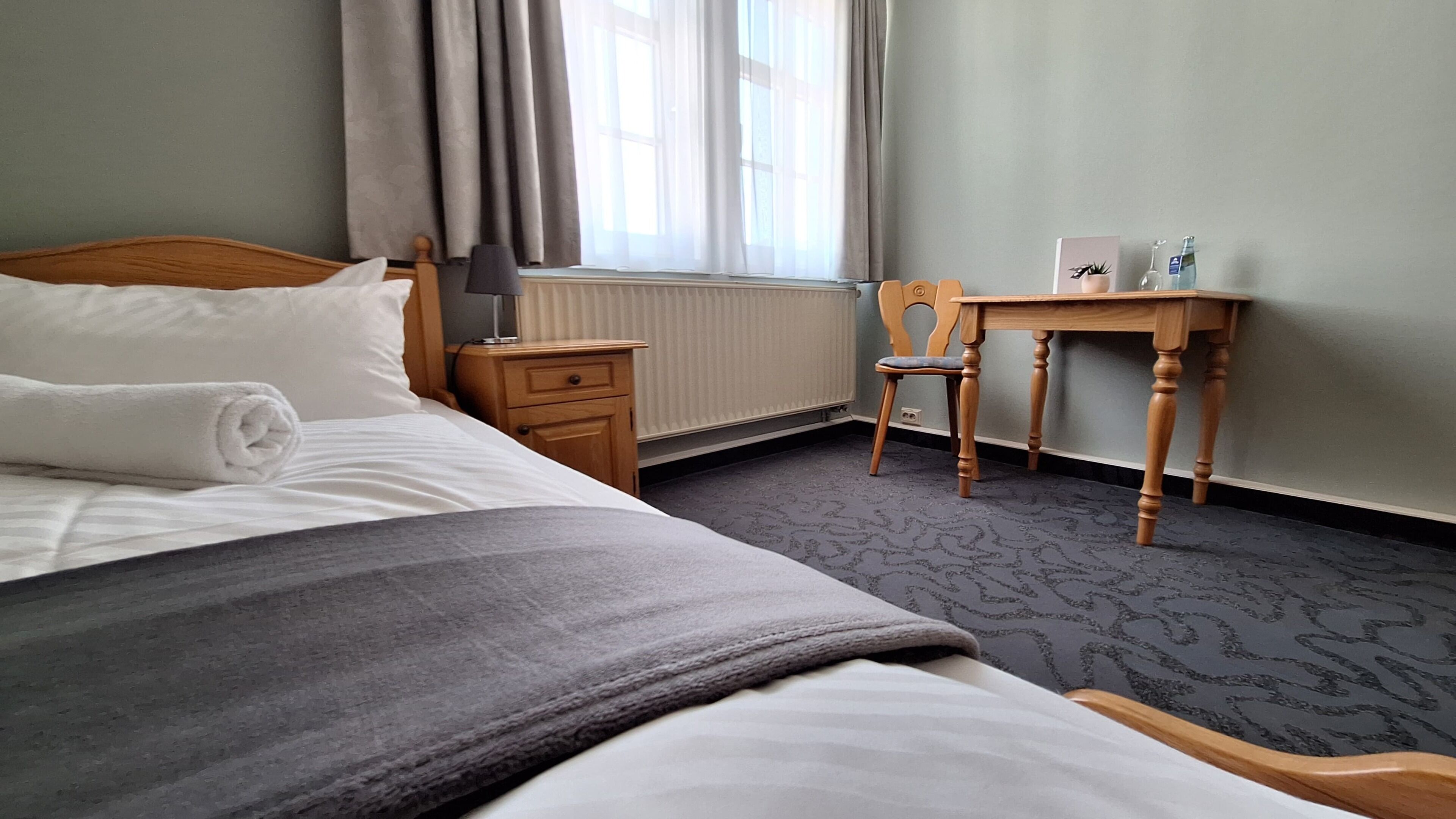 A hotel room with a bed, a desk, a chair, a nightstand, and a window with curtains.