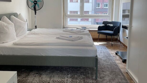A bedroom with a bed, a standing fan, a chair, a window, and a view of buildings outside. - Practical Studio nearby Lake (Cross 6) (Zürich)