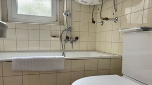 Een badkamer met een wit toilet, een bad met douchekop en een raam boven de wastafel.