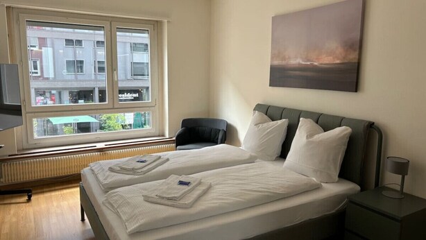 Hotelkamer met een bed, een stoel, nachtkastje, lamp en een raam met uitzicht op gebouwen.