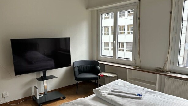 Une chambre d’hôtel avec un lit, une télévision, une chaise et une fenêtre donnant sur des immeubles.