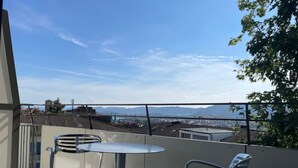 Un balcón con una mesa y dos sillas, con vistas a un paisaje urbano que incluye edificios y montañas a lo lejos.