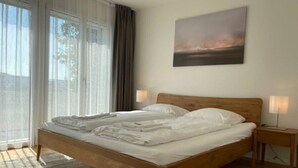 Un dormitorio con cama de madera, un ventanal con cortinas, un cuadro en la pared y una mesita de noche con lámpara.