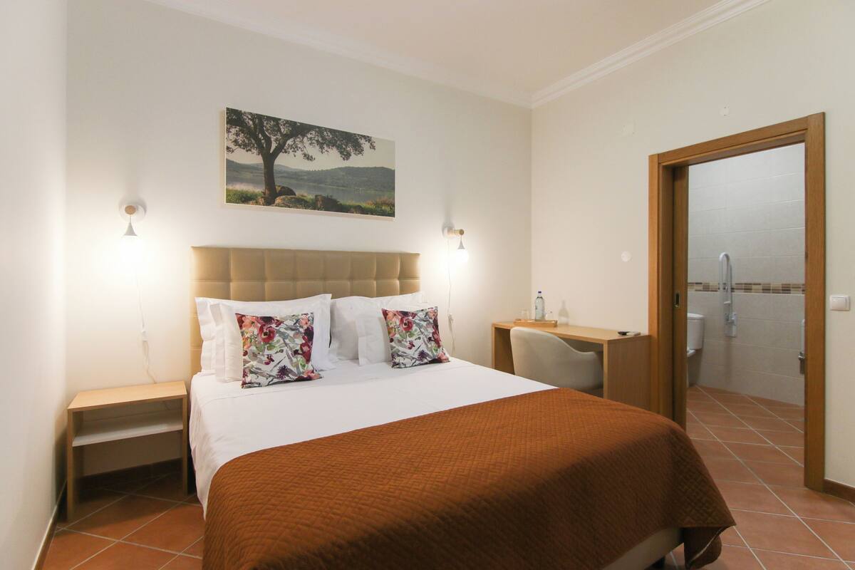 Ein Hotelzimmer mit Bett, Schreibtisch, Stuhl, Nachttisch, einem Gemälde und einem durchgegangenen Tür erkennbaren Badezimmer.